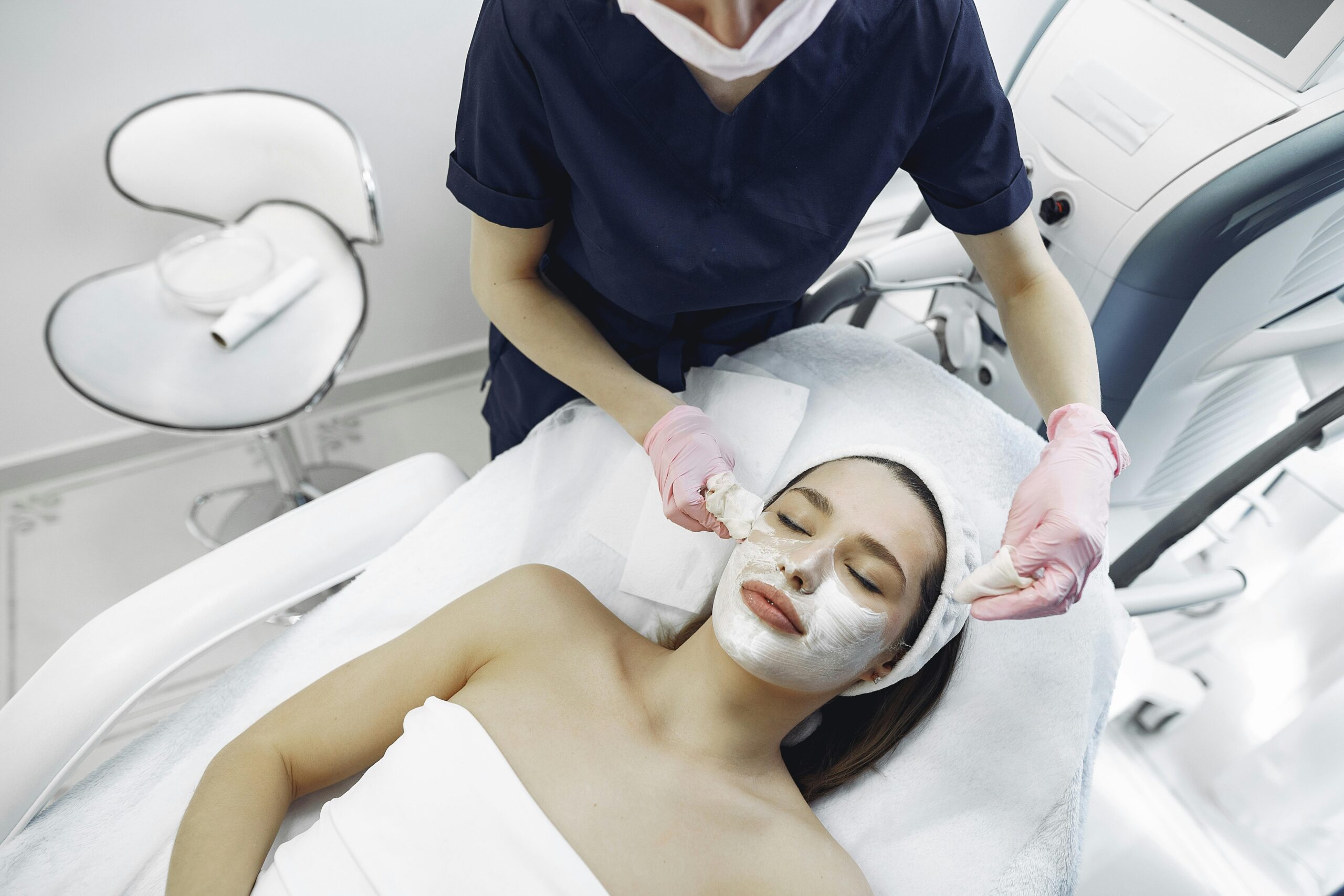 From above of dermatologist in medical coat and latex gloves doing face cleansing and wiping mask from face of female lying in towel in modern salon